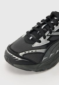 Schwarze Sneaker mit einem strukturierten Mesh-Obermaterial, silbernen Akzenten, dickem Gummisohle und einem kontrastierenden Muster an der Seite. Mit schwarzen Schnürsenkeln.