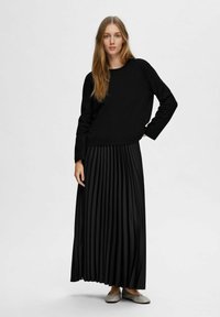 Schwarzes Langarmoberteil kombiniert mit einem schwarzen plissierten Maxirock. Glatter Stoff, schlichtes Design und ein passendes Paar hellgraue Schuhe.