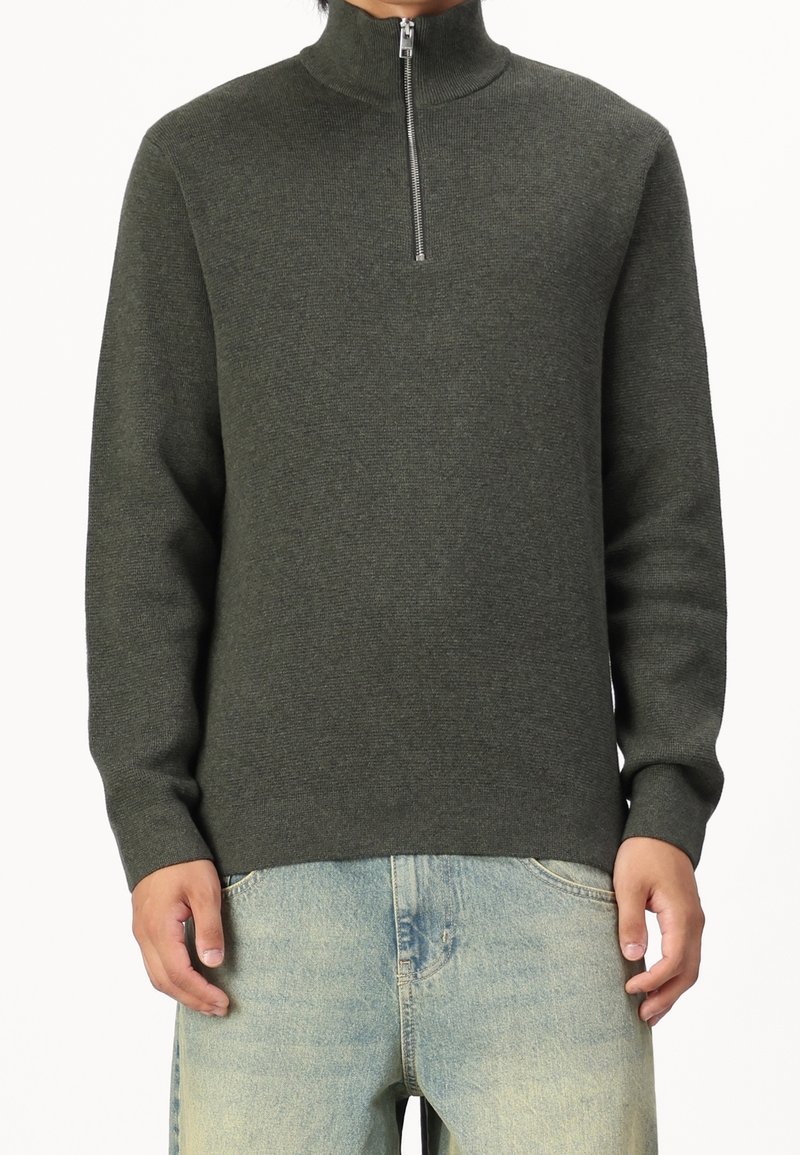 Homme portant un pull à demi-fermeture éclair vert foncé et un jean bleu clair délavé, debout les bras détendus le long du corps sur un fond blanc.