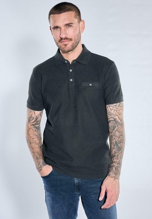 Man with short hair and tattoos on both arms wears dark polo shirt and blue jeans, standing with one hand in pocket against plain background.