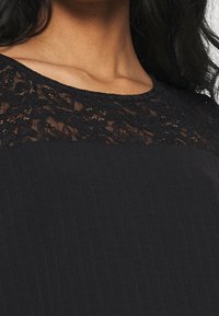 Haut noir avec un col en dentelle, tissu texturé et finition lisse. La dentelle présente un motif floral.