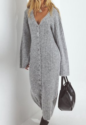 Kobieta ubrana w długą, szarą, prążkowaną sukienkę na guziki z szerokimi rękawami, trzymająca czarną skórzaną torebkę na białym tle.