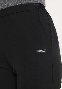 Černé sportovní kalhoty vyrobené z elastické tkaniny, s hladkým povrchem a malým bílým logem "VENICE BEACH" na boku.