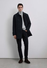 Abrigo de lana oscuro con cuello alto, usado sobre un jersey de cuello alto gris claro, combinado con jeans negros y botas de tobillo marrones. Diseño simple y ajustado.