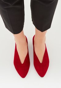 Rote Wildleder-Ballerinas mit spitzem Zehenbereich und tiefem V-Ausschnitt, kombiniert mit schwarzen, knöchellangen Hosen auf weißem Hintergrund. Weiche Textur und elegantes Design.