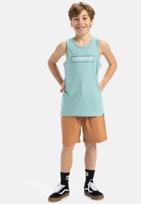 Ljusblå bomullslinne med en grafisk logotyp, kombinerat med orange shorts och svarta sneakers med vita detaljer samt mörka strumpor.