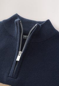 Laivastonsininen vetoketjullinen neule, jossa on resoripinta ja ribbineuloskaulus. Varustettu hopeisella vetoketjulla ja sileällä pinnalla. Etiketissä teksti: "NEX."