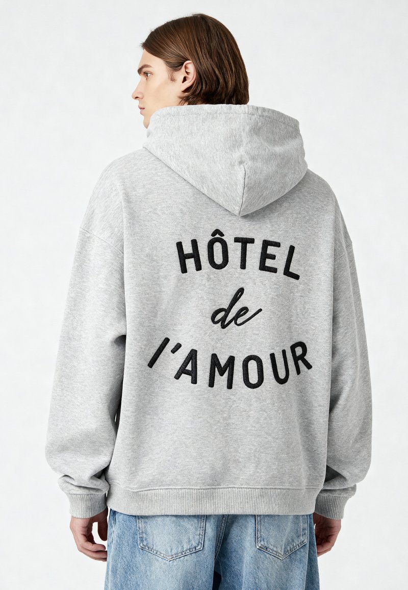 Szürke kapucnis felső, laza szabással, fekete hímzett felirattal a hátsó részén, amely így szól: "HÔTEL de l'AMOUR", valamint bordázott mandzsettákkal.