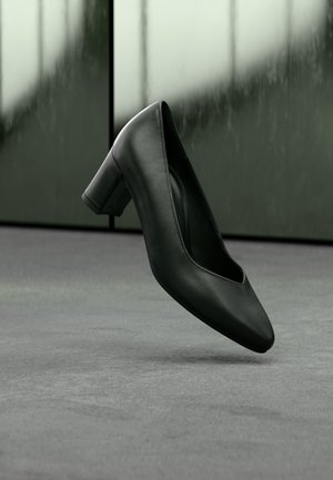 Schwarzer Damenlederschuh mit niedrigem Blockabsatz, der über einer grauen Oberfläche mit verschwommenem grünem und schwarzem Hintergrund schwebt.