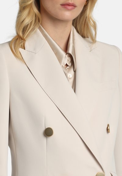 Blazer beige croisé avec boutons dorés, revers ajustés et texture lisse ; associé à une chemise en satin claire.