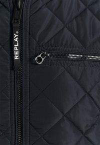 Quilted black jacket fabric with diagonal stitching. Features a central zipper and a small zippered pocket on the left side. Branding visible.