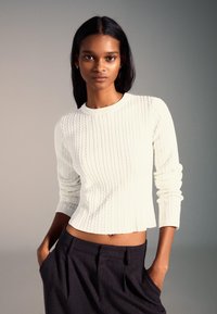 Biały prążkowany sweter z okrągłym dekoltem i dopasowanymi rękawami, w połączeniu z ciemnymi, eleganckimi spodniami. Gładka faktura i krótki krój.
