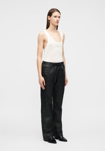 Γυναίκα που φοράει λευκό τοπ χωρίς μανίκια με στρογγυλή λαιμόκοψη, χαλαρό μαύρο παντελόνι με υφή και μαύρα ψηλοτάκουνες μυτερές γόβες, στέκεται μπροστά σε απλό φόντο.