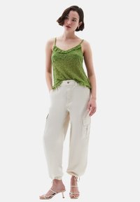 Zelený strukturovaný top na tenkých ramínkách vyniká v kombinaci s krémovými cargo kalhotami se bočními kapsami a elastickými manžetami. Béžové sandály na podpatku.