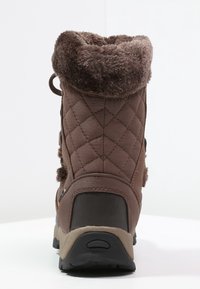 Bota de invierno marrón con exterior de tejido acolchado, cuello de piel sintética, suela de goma resistente, cremallera lateral y fondo texturizado para mayor agarre.