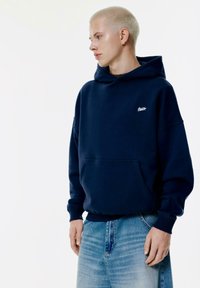 Felpa navy in un materiale morbido con tasca a marsupio frontale, polsini a coste e un piccolo logo bianco sul petto. Abbinata a jeans azzurri chiari.