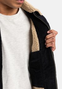 Navy corduroyjakke med en beige fleeceforing, der har en lynlåslukning og en struktureret overflade. Båret over en lys grå skjorte.