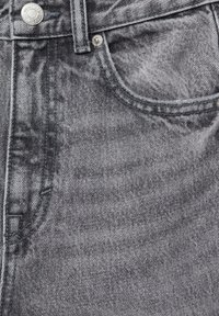 Primer plano de un bolsillo de vaqueros de mezclilla gris con costuras visibles, botón de metal y remache sobre una tela texturizada.
