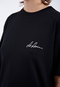 Camiseta de algodón negra con cuello redondo, que presenta una firma bordada blanca en la parte izquierda del pecho. Corte clásico, mangas cortas.