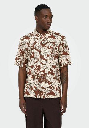 Homme portant une chemise à manches courtes marron et beige avec un grand motif de feuilles tropicales, regardant vers sa gauche sur un fond uni.