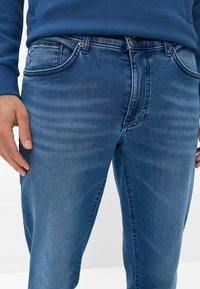 Jeans in denim blu con taglio dritto, caratterizzati da una leggera scoloritura, design a cinque tasche, bottoni metallici e dettagli di cucitura ben visibili.