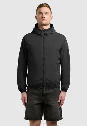 Mężczyzna w czarnej kurtce z kapturem na suwak i ciemnoszarych denimowych szortach, stojący na tle jednolitego, jasnego tła.