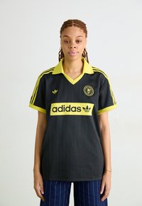 Melna Adidas krekls ar dzelteniem akcentiem, ar apkakli, trijām svītrām uz piedurknēm un lielo logotipu uz priekšpuses. Vertikālas svītras uz auduma.