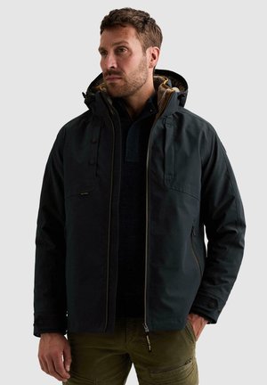 Homme aux cheveux courts et à la barbe portant une veste noire à capuche par-dessus un pull sombre et un pantalon vert olive avec des poches.