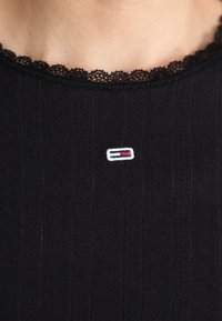 Svart topp med blonderkant i halsen, med en liten brodert logo i hvit, rød og blå. Stoffet har en ribbet tekstur.