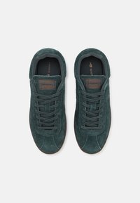 Green suede sneakers with tonal laces, round toe, and textured surface. Features a brown brand label on the tongue. Rubber sole.