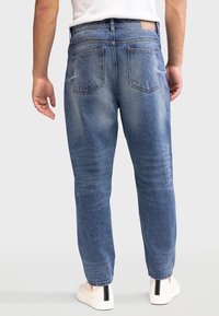 Jean en denim bleu avec une coupe décontractée, doté de deux poches arrière et d'une jambe légèrement fuselée. Porté avec des baskets blanches.