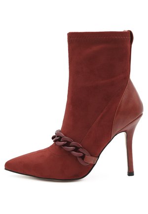 Stivale alla caviglia in suede rosso con punta appuntita e alto tacco a stiletto. Presenta un accento di catena spessa attorno alla caviglia. Texture liscia.