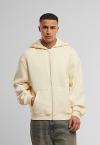 Cremefarvet zip-op hoodie lavet af blødt stof, med en rummelig hætte, frontlommer og ribbede manchetter, parret med denim jeans.