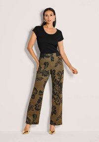 Vrouw in een zwart aansluitend T-shirt met korte mouwen, hooggetailleerde bruine broek met grote zwarte bloemenpatronen en gouden puntige hoge hakken.