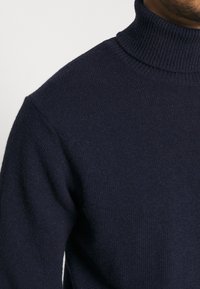 Maglione a collo alto a coste blu navy scuro indossato da una persona, che mostra la parte superiore del busto e l'area del collo.