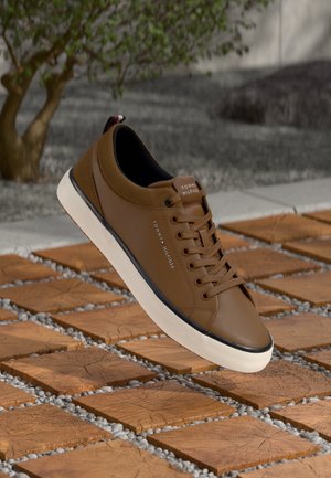 Brązowy skórzany sneaker Tommy Hilfiger z białą podeszwą, czarną lamówką i sznurowadłami, prezentowany na drewnianej i kamiennej podłodze na zewnątrz.