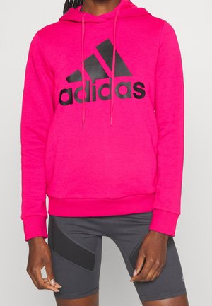 Person wearing bright pink Adidas hoodie with black logo and dark gray athletic shorts with black panels.