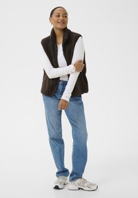 Mujer con camisa blanca de manga larga, chaleco de forro polar marrón oscuro, vaqueros azules y zapatillas blancas, de pie con los brazos cruzados y sonriendo.