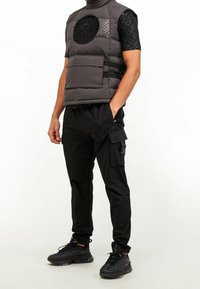 Gilet imbottito grigio con un logo nero rotondo, abbinato a pantaloni cargo neri e sneaker nere. Il gilet presenta un collo alto e tasche frontali.