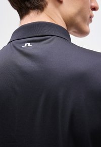 Άνδρας που φοράει σκούρο πόλο πουκάμισο, δείχνοντας το πίσω μέρος του γιακά με ένα μικρό λευκό λογότυπο στο κέντρο κάτω από το λαιμό.