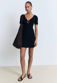 Black short-sleeve dress with a v-neck and drawstring detail, paired with a large brown woven bag and black flat sandals.