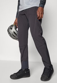 Dunkelgraue Outdoor-Hose mit verstärkten Knien, Reißverschlusstaschen und glatter Textur, kombiniert mit schwarzen Turnschuhen und einem Helm.