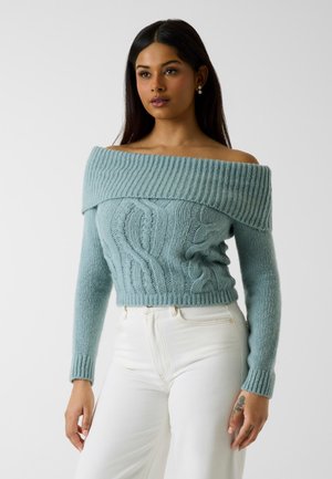 Giovane donna che indossa un maglione azzurro chiaro lavorato a maglia con motivi a trecce e pantaloni bianchi a vita alta, in piedi su uno sfondo semplice.