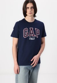 Mornarski pamukasti T-shirt s hrabrim natpisom "GAP ITALY" u bijeloj i crvenoj boji. Ima okrugli izrez i kratke rukave, nosi se uz svijetlo plave traperice.