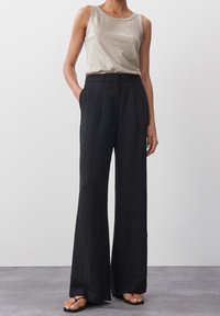 Mustat, leveälahkeiset housut, joissa on pleat't ja sivutaskut, yhdistettynä kiiltävään, vaaleanharmaaseen hihattomaan puseroon. Litteät sandaalit täydentävät asua.