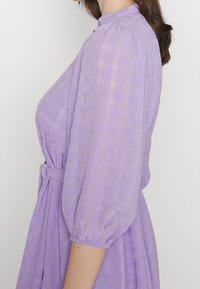 Lavendelfarbenes, strukturiertes Kleid mit hohem Kragen, gepufften Ärmeln und gebundenem Taillenband. Der Stoff weist ein Rautenmuster für zusätzliche Details auf.