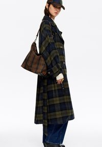 Karirasti dolg plašč temno zelene in mornarske barve, z velikimi gumbi, zanka za obešanje in bočnim razporkom. V kombinaciji z vzorčasto torbico in kavbojskimi hlačami.