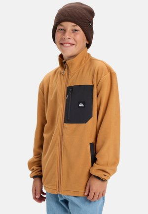 Veste en polaire beige avec une poche poitrine noire et un logo, dotée d'un col haut et d'une fermeture éclair. Portée avec un bonnet en tricot marron.