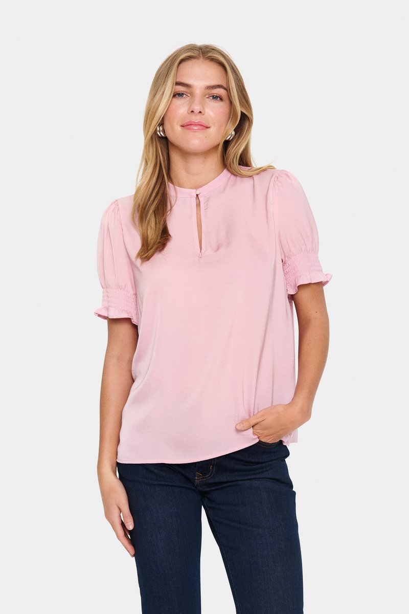 Blusa rosa chiaro con scollatura a chiave, maniche corte a sbuffo con dettagli arricciati e vestibilità comoda, abbinata a jeans blu scuro.