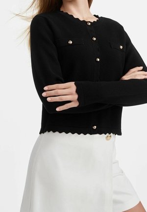 Femme portant un cardigan noir boutonné avec des bords festonnés et une jupe blanche avec un bouton doré, bras croisés sur la poitrine.